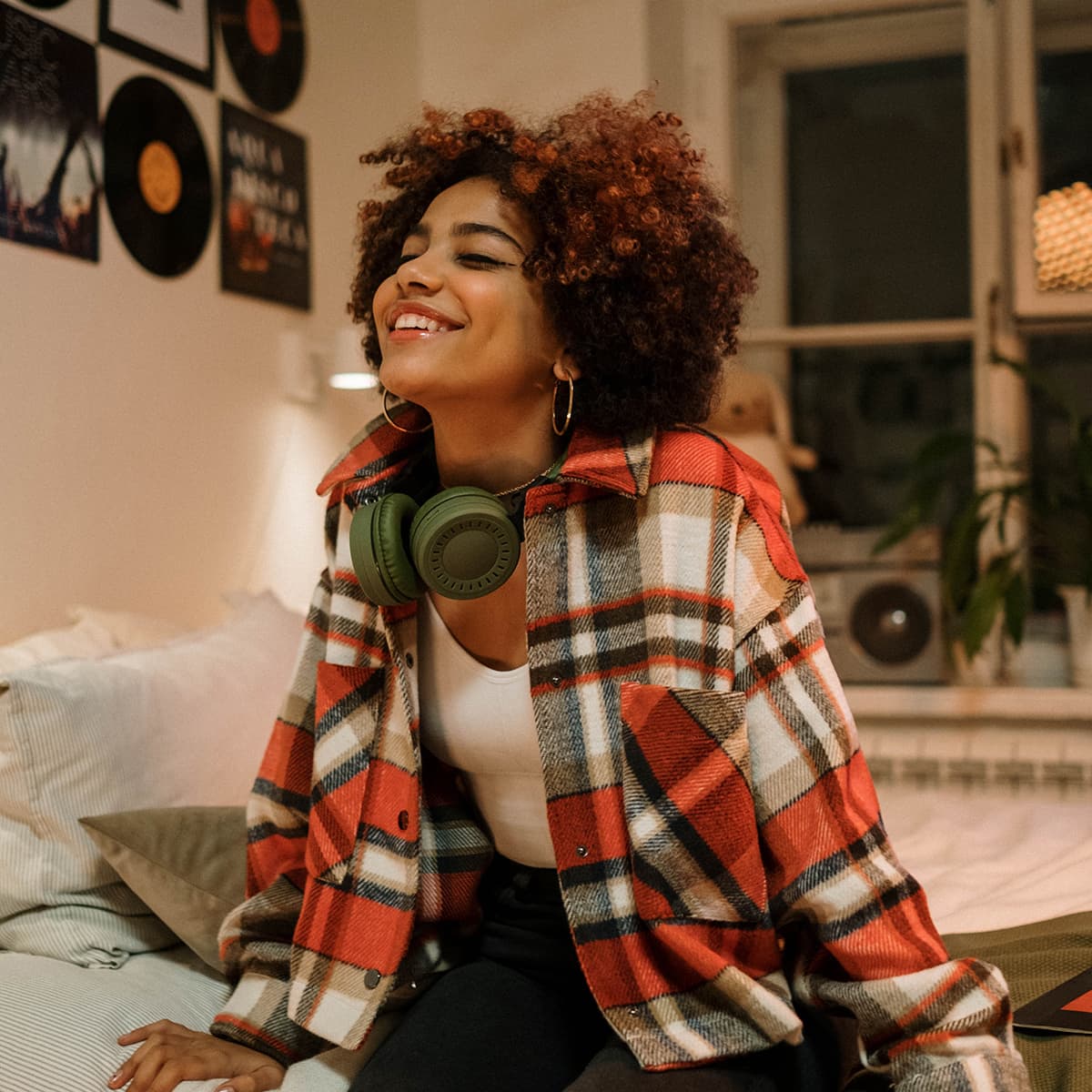 A Black person with curly red hair sitting in a bed smiling with headphones around their neck