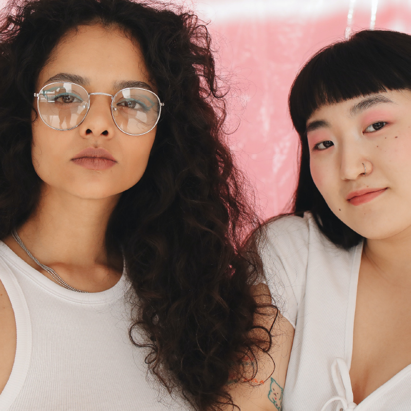A close up shot of two young people who gaze at the camera. One has light brown skin with long wavy hair and glasses. She wears a white tank top and a serious expression. The second person has shoulder-length black hair with straight bangs. She has a slight smile and wears a v-neck t-shirt.