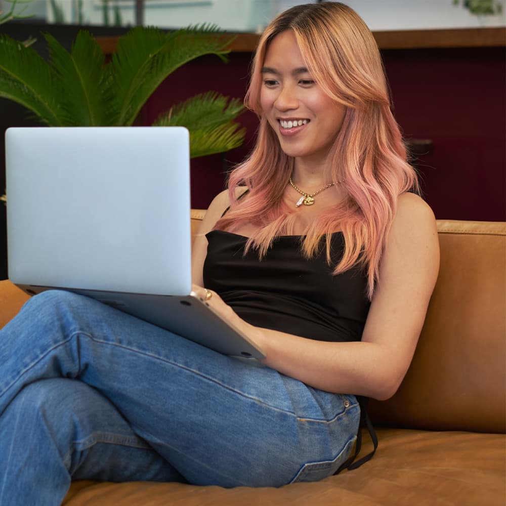 A young person with pink and blonde hair, sitting and smiling on a brown couch while looking at a laptop screen.