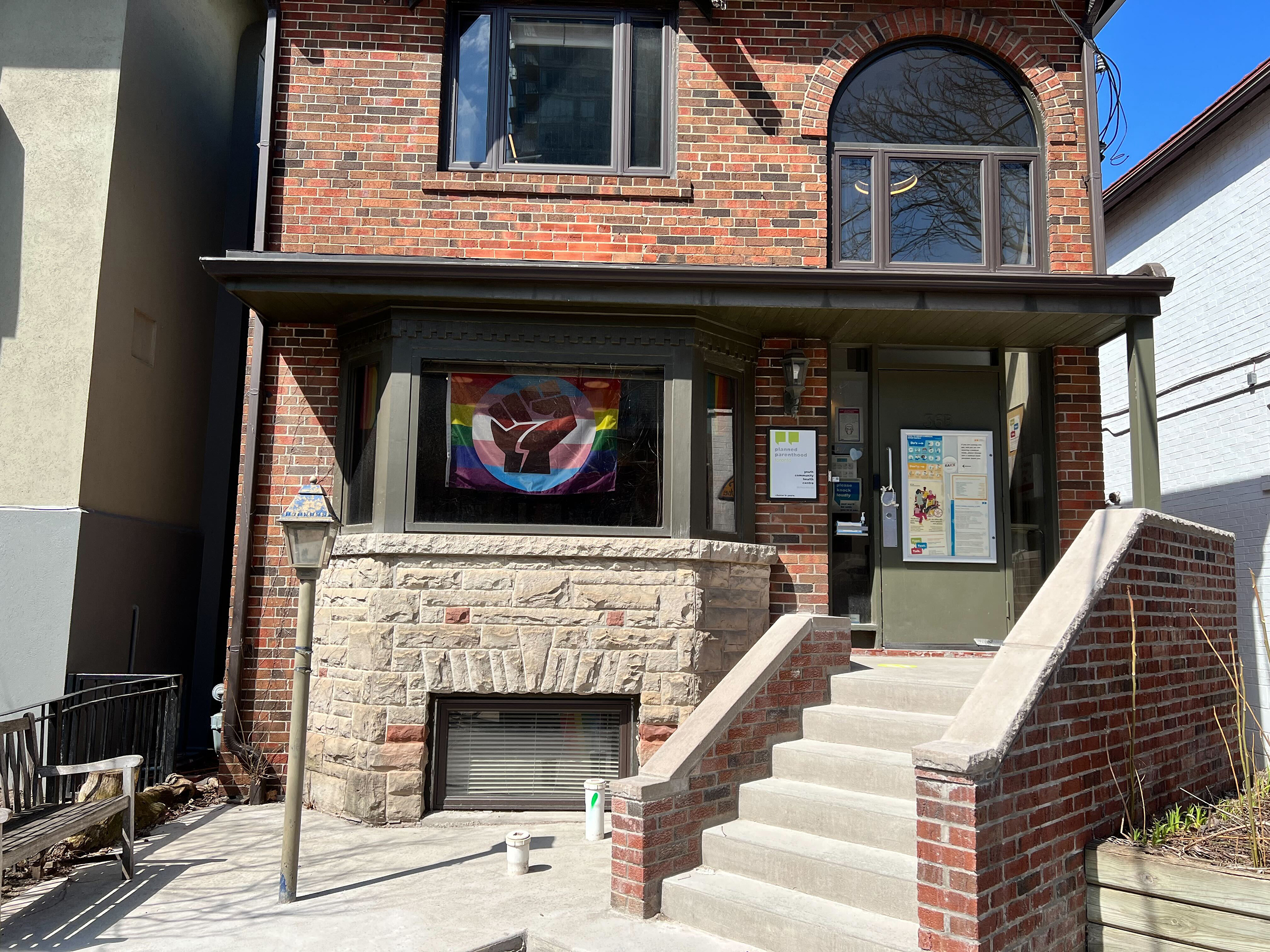 The PPT Headquarters on a sunny day: a brick house with an arched window and a Pride Power flag in the front window.