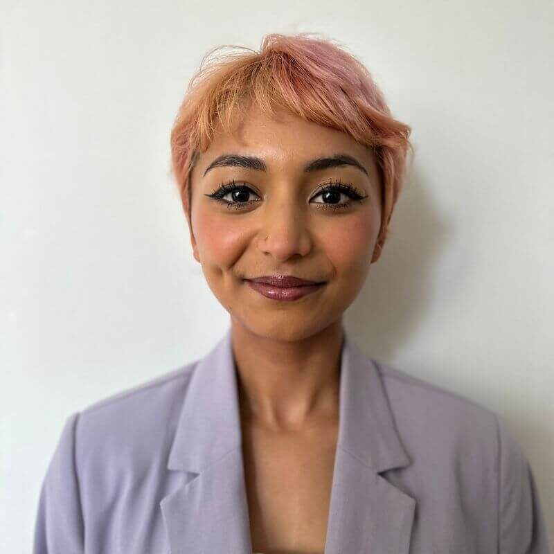 A headshot of Ashmita, smiling into the camera with short pink hair, wearing a purple blazer.