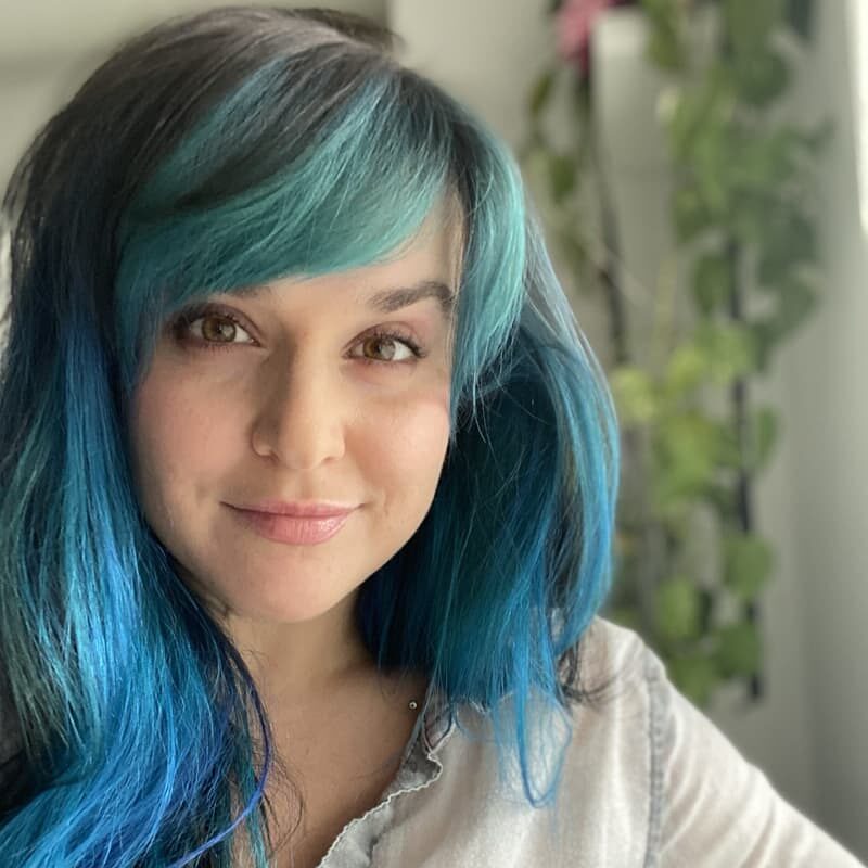 A headshot of Olga smiling at the camera with blue ombre hair and a hanging plant in the background