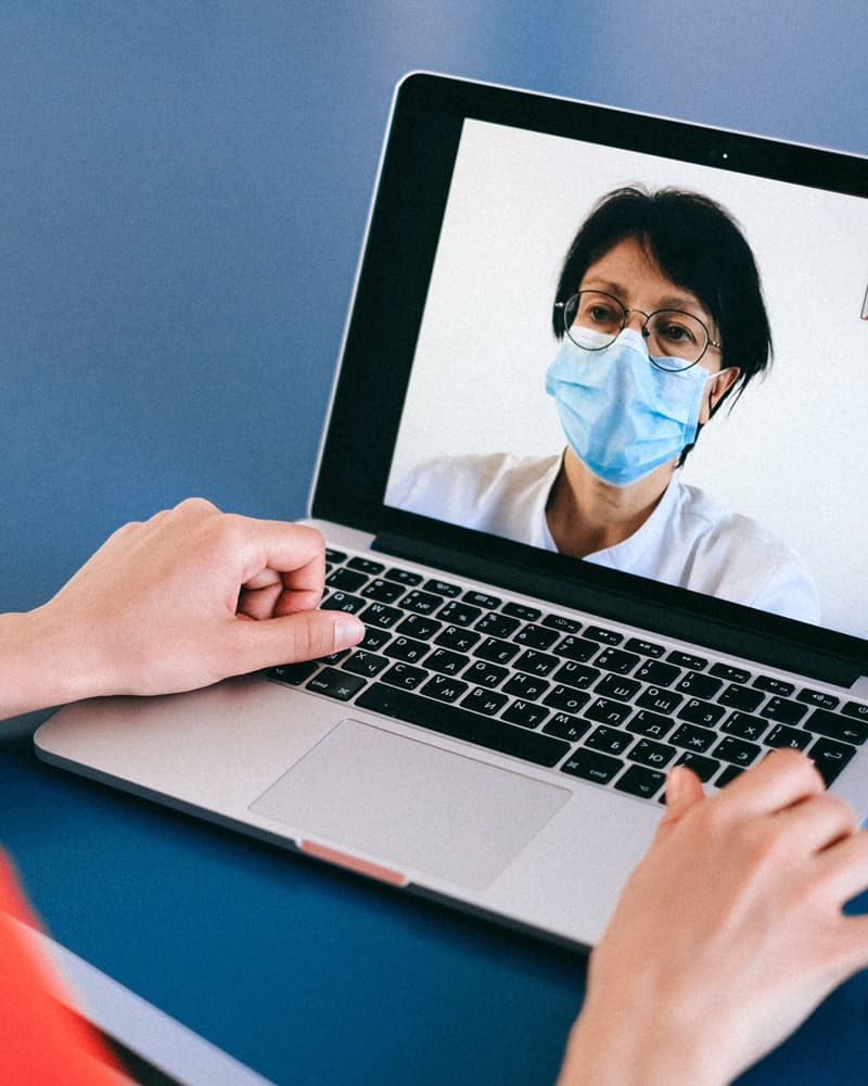 A laptop screen, where a medical professional wearing a face mask is consulting digitally with a patient.