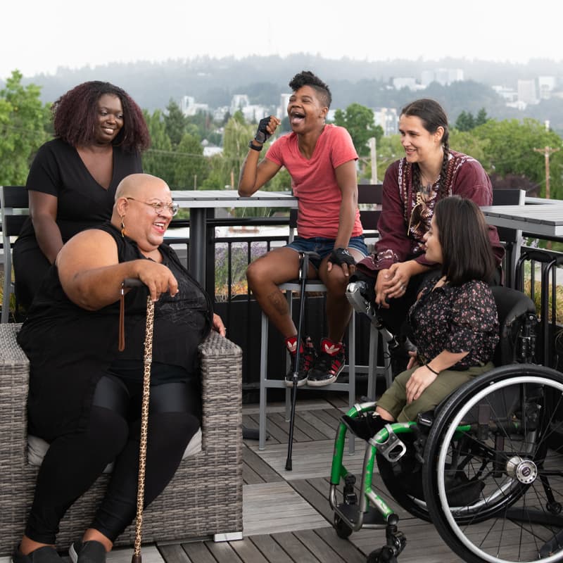 Five disabled people of color with canes, prosthetic legs, and a wheelchair sit on a rooftop deck, laughing and sharing stories. Greenery and city high-rises are visible in the background.