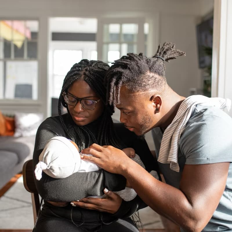 A young, Black couple holds a baby. The mother has glasses and long braids. The father has short dreadlocks in a ponytail with an undercut. They are seated in chairs in their living room, which is bright and well lit. They are looking at the baby with a calm, loving expression.