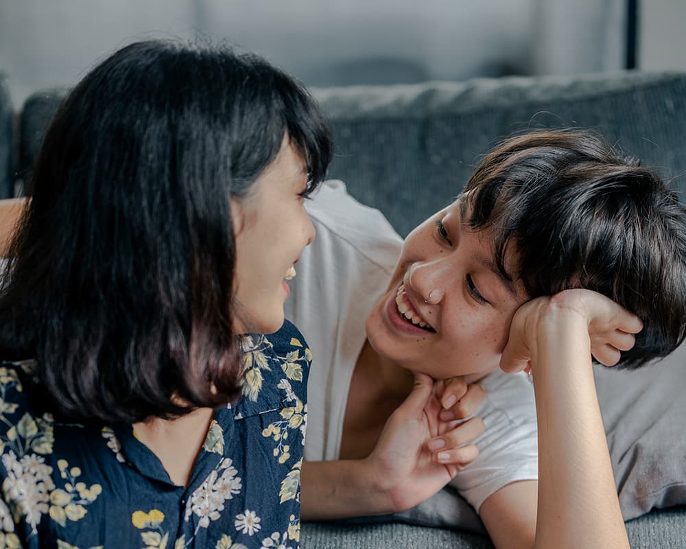 Two folks of Asian decent, one with short hair and a nose ring, and the other with longer hair and a floral shirt, looking at eachother lovingly and smiling on a couch.