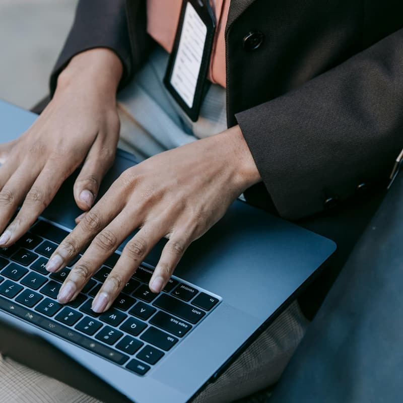 A zoomed in shot of a person working on a laptop. The frame shows the person’s torso and the laptop sitting on their lap. The person has brown skin, and is wearing a black blazer, coral shirt, ID lanyard badge, and grey dress pants.