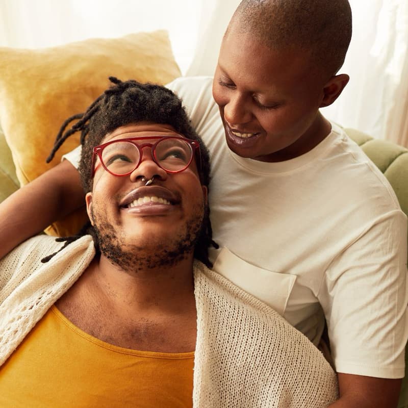 Two young Black folks, one with a yellow shirt and bright red glasses, and the other with white t-shirt and shaved head, cuddling on a green couch and smiling at one another.