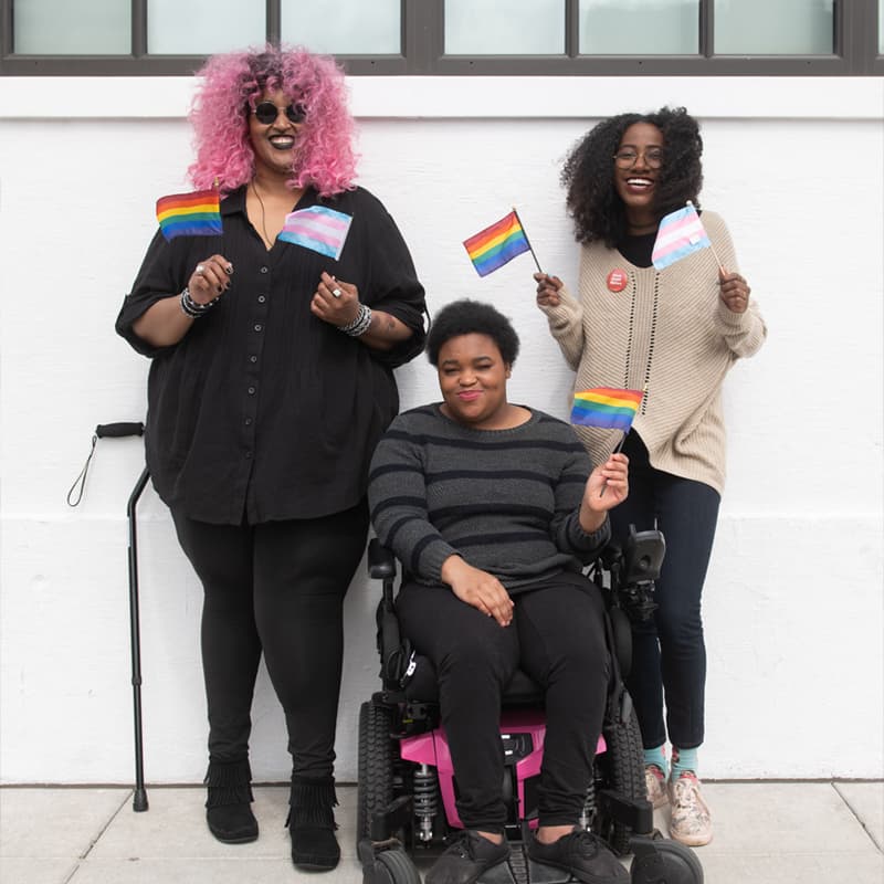 Three Black and disabled folks smile and hold mini flags. On the left, a non-binary person holds both a rainbow pride flag and a transgender pride flag, while a cane rests behind her. In the middle, a non-binary person waves the rainbow flag while in their power wheelchair. On the right, a femme waves both a rainbow and transgender pride flag.