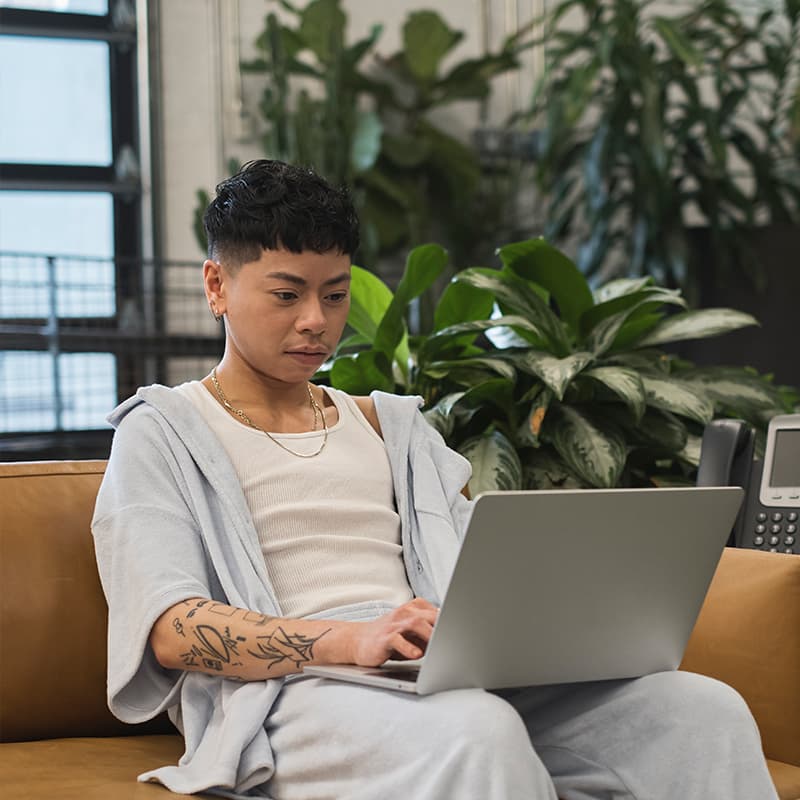 A non-binary person sits on a couch using a laptop. The room has a converted warehouse/industrial vibe. Light streams in through a window, and multiple tropical plants fill the background.