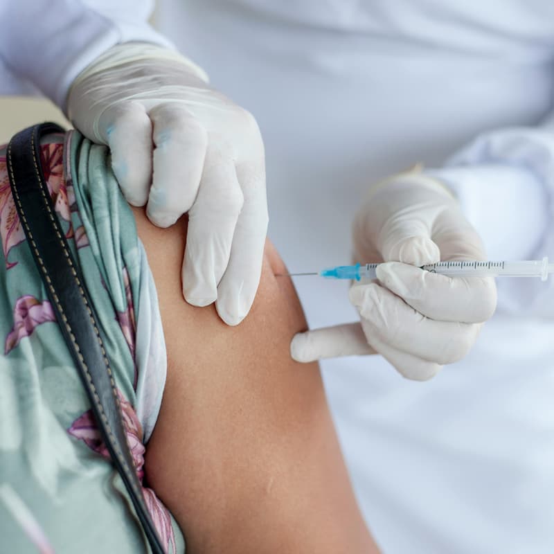A zoomed-in shot of a person’s upper arm receiving a vaccination. The person has light brown skin, and is wearing a flowered shirt and a mask. The person delivering the vaccine is wearing a white lab coat and gloves.