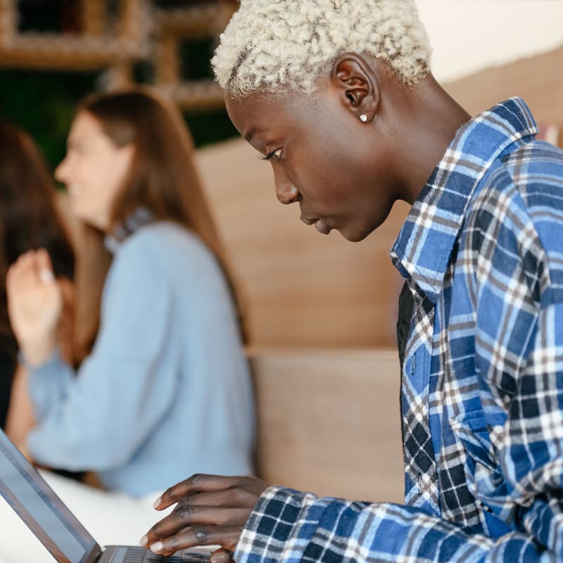 A young Black person with short blonde hair and studded earrings, wearing a plaid shirt and working on a laptop.