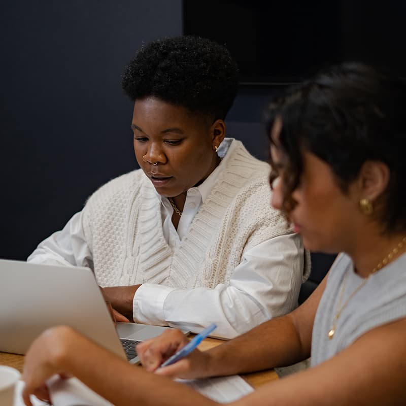 Two youth of colour sit at an office table, working. One has short, curly black hair, is wearing a white sweater vest, and works at a laptop. The other has longer black hair pulled back and is wearing gold jewellery and a grey tank top. This person is writing on a notepad.