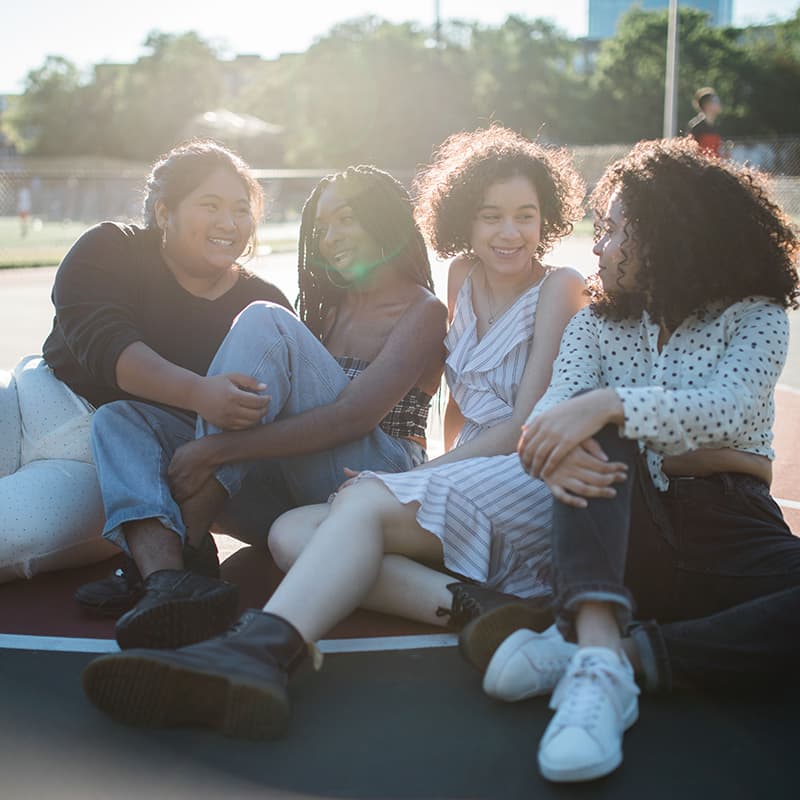 Four young people, representing a mix of genders and ethnicities, sitting and talking on a outdoor basketball court on a sunny day.