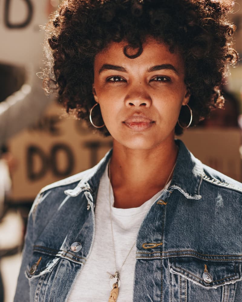 A young Black person with curly hair and small hoop earrings, lit by the sun, looking with a serious tone towards the camera.