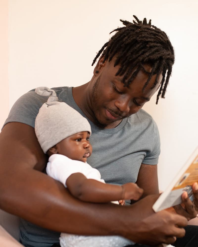 A young Black father reading a book to a baby that is cradled in his arms.