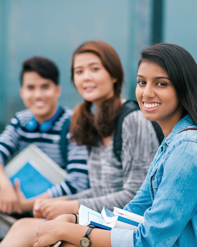 A diverse group of young folks, high school aged, holding books and smiling at the camera.