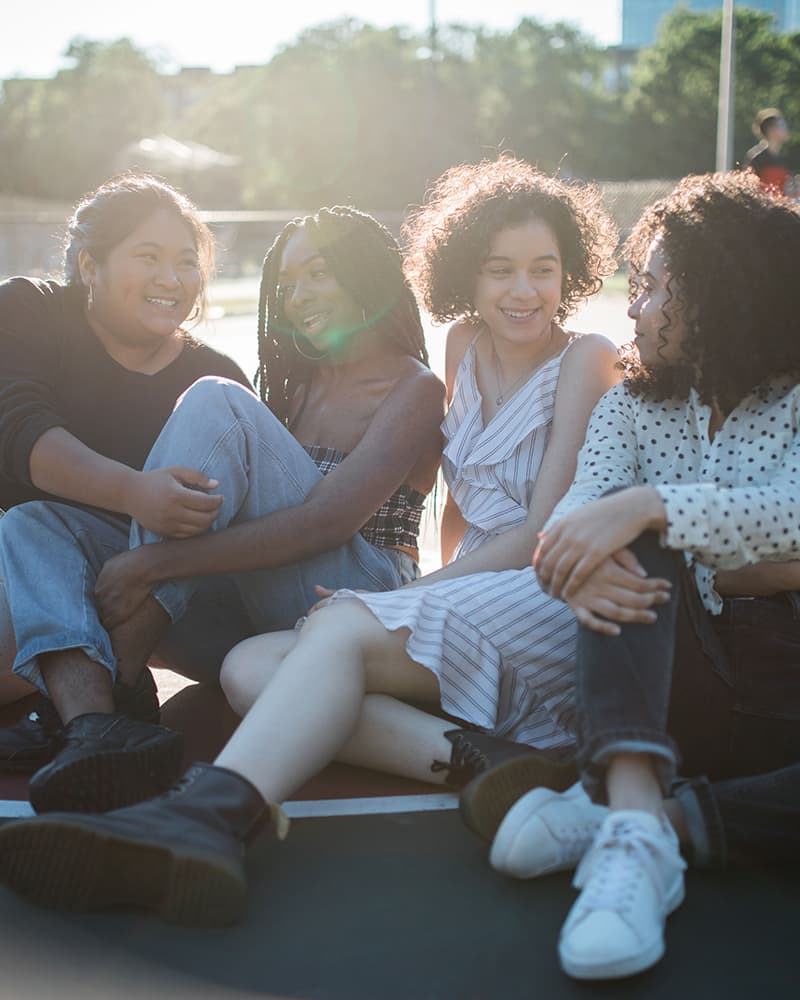 Four young people, representing a mix of genders and ethnicities, sitting and talking on a outdoor basketball court on a sunny day.